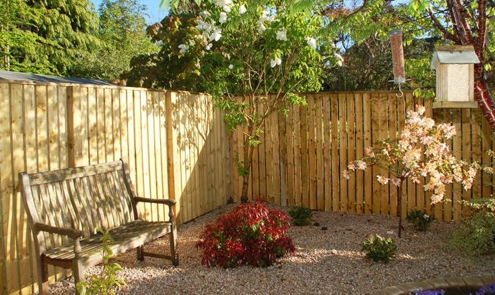Double boarded palisade timber fencing around a gravelled garden corner with a bench