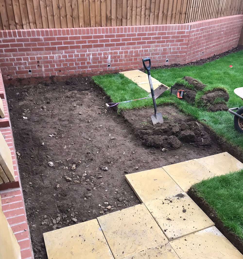 Turf lifted and ground dug out ready for paving slabs beside a brick wall